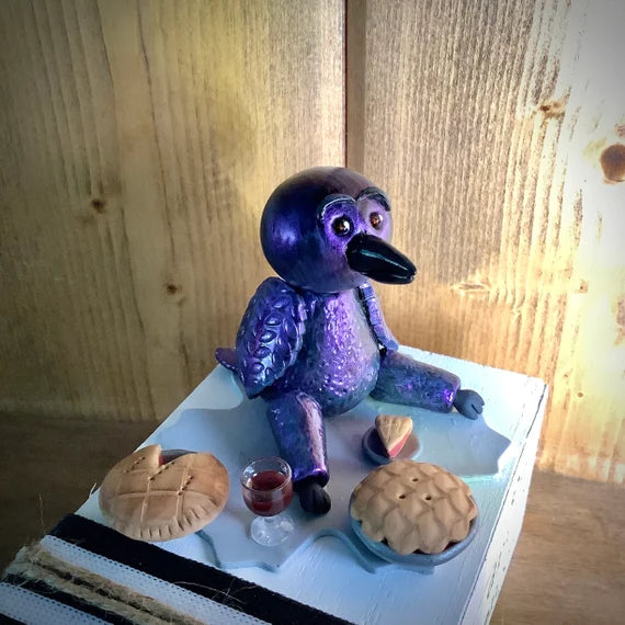 Eat Drink Enjoy Crow with Mini Glass of Red Wine and 2 Mini Pies, Black Purple Blue Color Shifting Clay Raven Sitting on White Book Stack